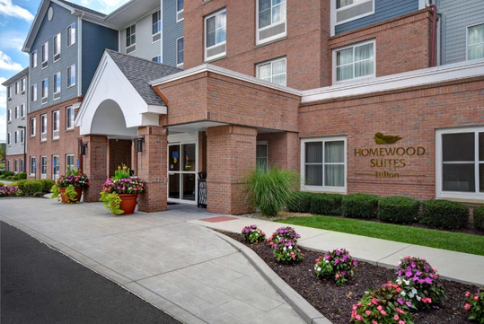 Exterior view of a Homewood Suites hotel by Hilton, featuring a brick facade, white arch entrance, and landscaped flower beds.