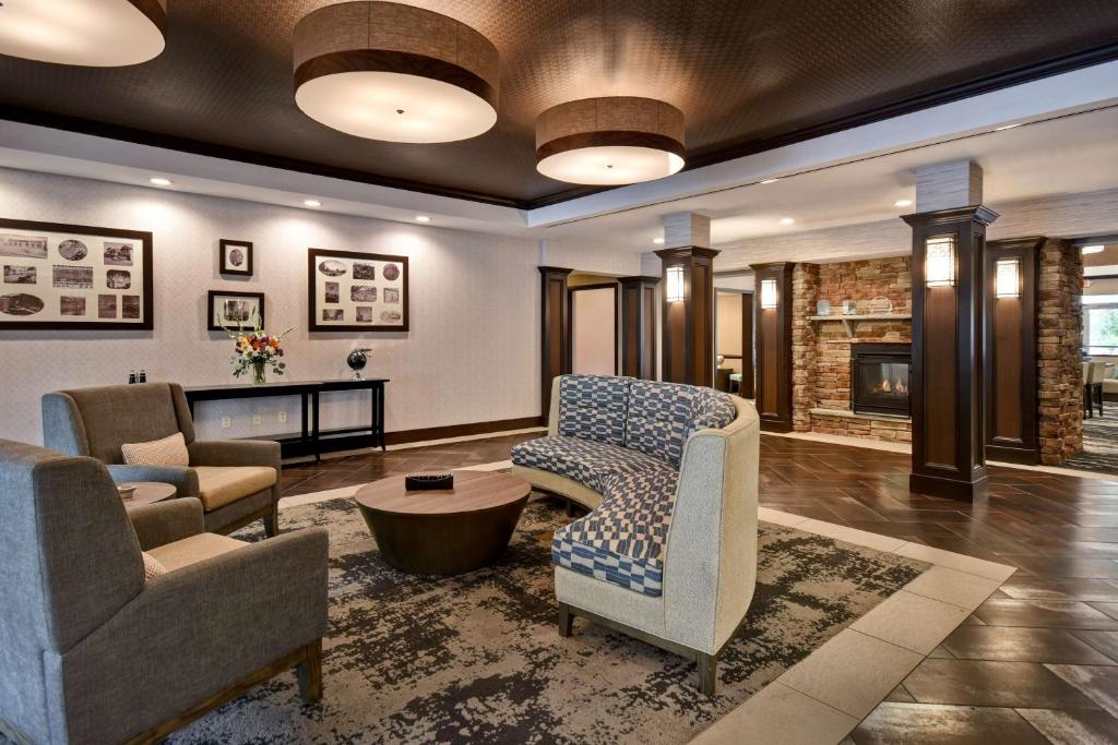Hotel lobby with modern decor. Features circular seating, coffee table, and ceiling lights. Brick fireplace and framed pictures on walls. Neutral tones and patterned rug on tiled floor.