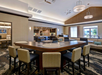 A modern hotel lounge with a central high table and chairs. The room has pendant lights, a patterned carpet, and large windows allowing natural light.