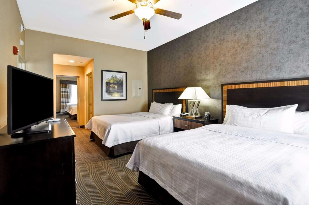 Hotel room with two double beds, a TV, and a ceiling fan. A lamp sits on the bedside table between the beds. There is a hallway leading to another part of the room.
