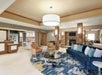 Spacious lobby with a modern design, featuring a blue couch, beige chairs, a round rug, and a stone fireplace. A large overhead light fixture and snack area are visible.
