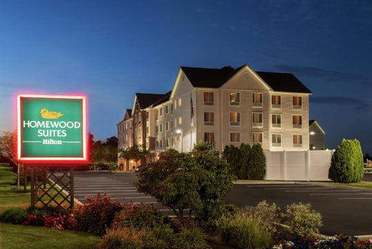 Four-story Homewood Suites hotel at dusk with illuminated sign in foreground and landscaped parking area.