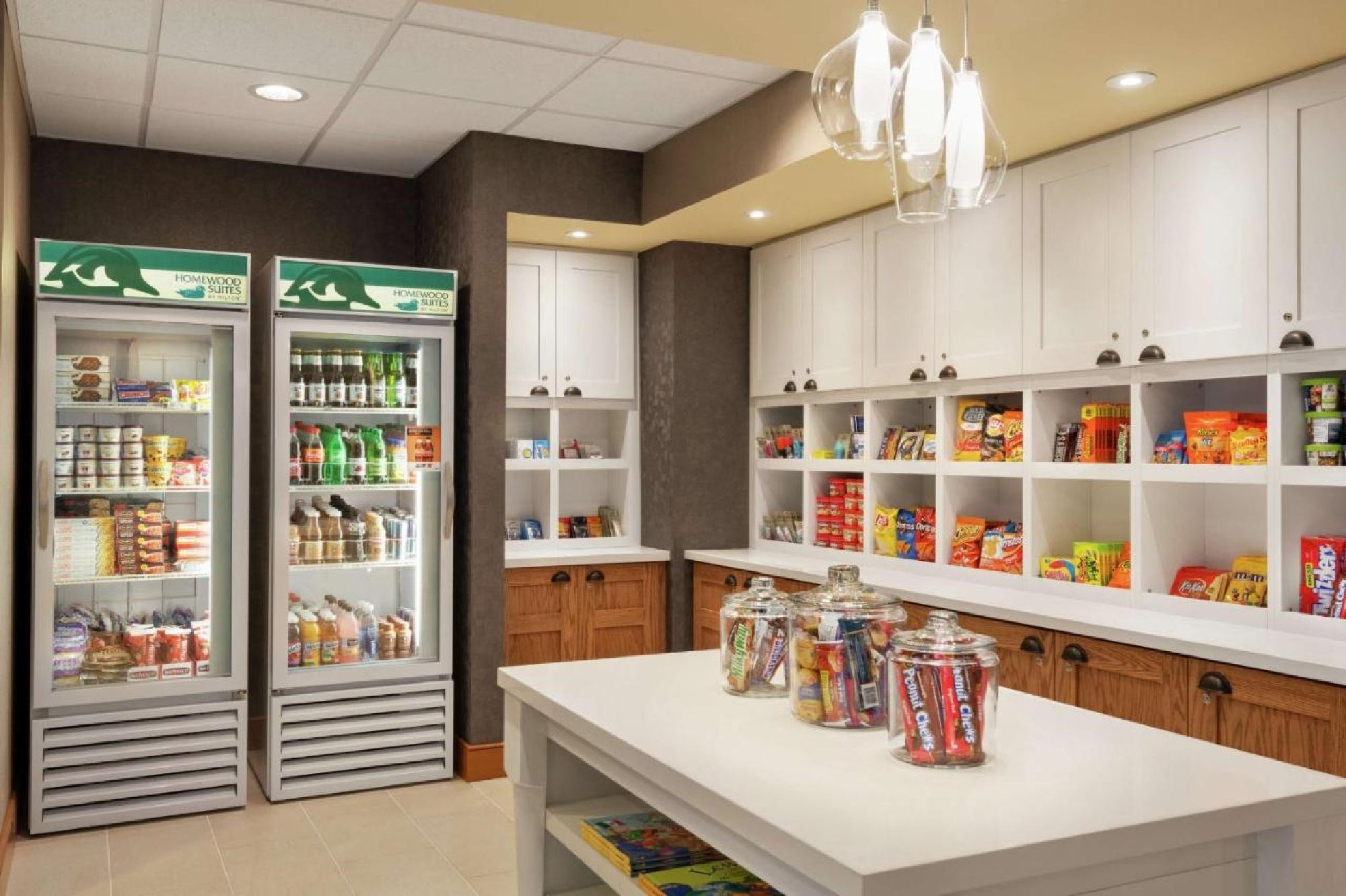 A small convenience store with two refrigerators and shelves filled with snacks, drinks, and various food items. A countertop displays glass jars containing candy.