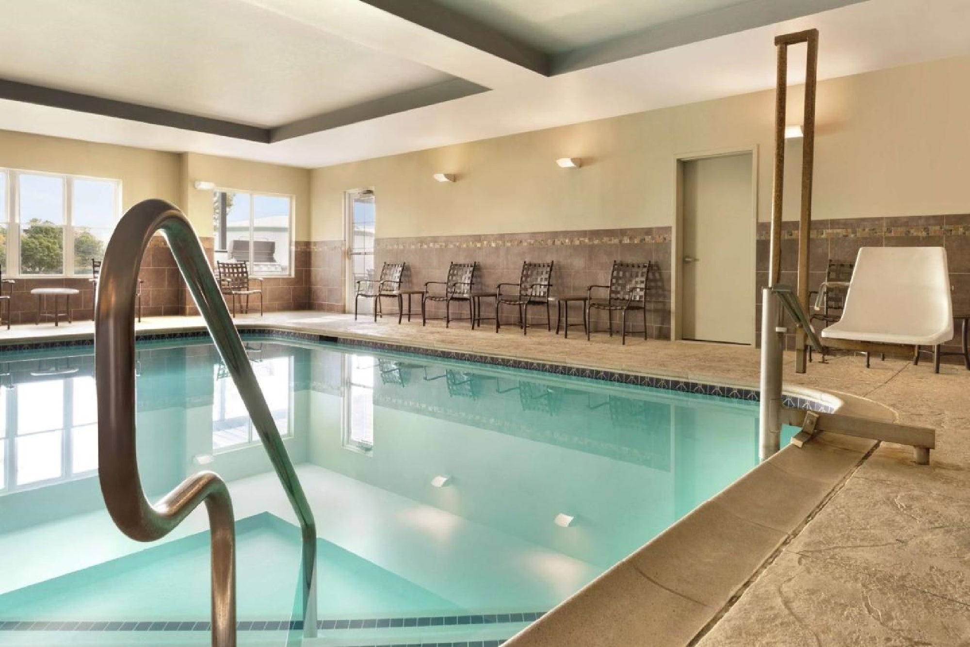 Indoor swimming pool area with chairs along the walls, windows on one side, and a pool lift in the corner.