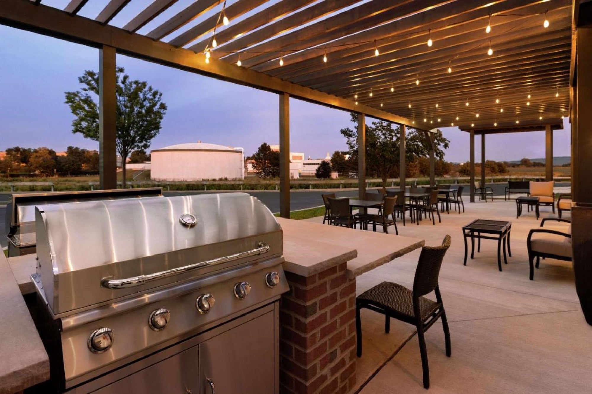 Outdoor patio with a grill, seating area, and string lights under a pergola. Grassy field and trees in the background during evening.