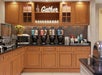 Coffee station with four dispensers, a microwave, cereal dispensers, and a fruit basket. Wooden cabinets and a sign reading "Gather" above.