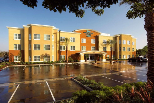 Front view of a three-story hotel building with a central entrance, surrounded by a wet parking lot and landscaped greenery.