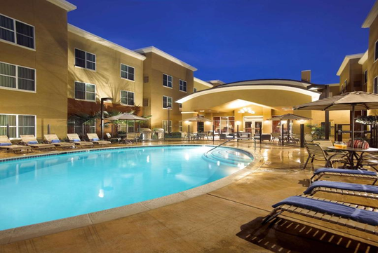 Outdoor hotel swimming pool with lounge chairs and umbrellas, surrounded by a multi-story building, lit up in the evening.