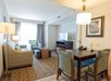 A hotel room with a sitting area includes a sofa, two chairs, a TV on a dresser, and a small dining table with two chairs. There's a lamp, overhead light, and a window with curtains.