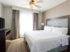 A hotel room featuring a neatly made bed, a wooden dresser with a TV, a floor lamp, and a window with curtains. A ceiling fan is above the bed.