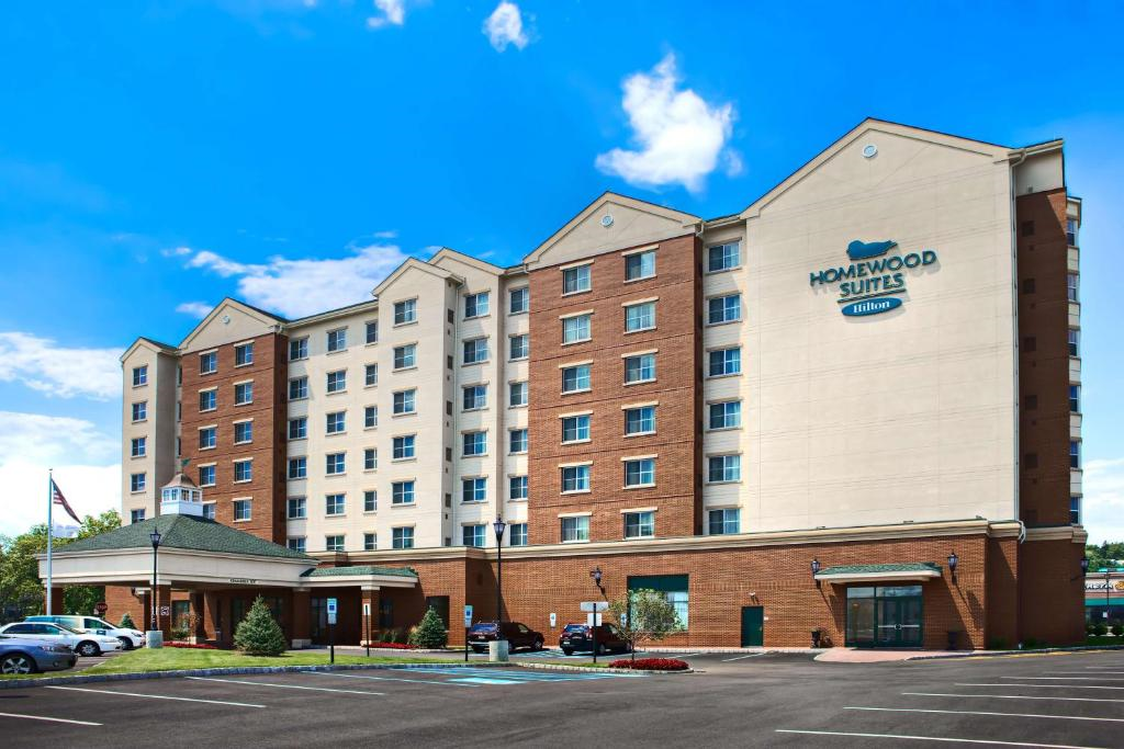 Exterior view of a multi-story Homewood Suites by Hilton hotel with brick and cream facade, main entrance, parking lot, and cars under a partly cloudy sky.