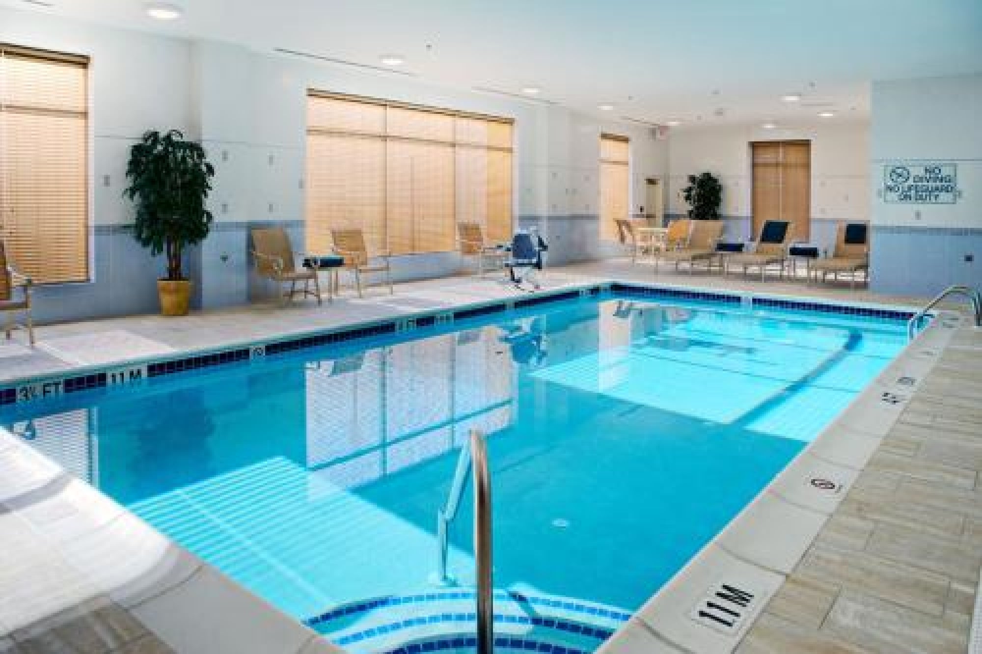 Indoor swimming pool with clear water, surrounded by chairs, tables, and potted plants; large windows with blinds let in natural light.