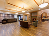 Spacious hotel lobby with wooden flooring, a stone fireplace, sofas, armchairs, and a reception desk. Chandeliers hang from the ceiling, and there is a dining area in the background.