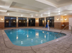 Indoor swimming pool with a hoist, surrounded by a tiled deck and illuminated by wall lights. Large windows line the far wall.