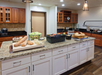A kitchen with a granite island displays bread, salad, cheese, and other food items. Wooden cabinets and a coffee maker are in the background.