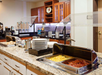 Buffet-style breakfast area with trays of scrambled eggs and bacon on a granite counter, alongside plates, utensils, and cereal dispensers in the background.