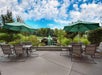 Patio area with two tables and green umbrellas, surrounded by chairs, overlooking a small fountain and landscaped greenery under a partly cloudy sky.