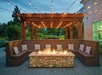 Outdoor seating area with brown wooden benches, patterned pillows, a stone fire pit, and a wooden pergola overhead with string lights, surrounded by greenery.