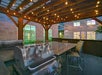 Outdoor kitchen area with a stainless steel grill, bar seating, stone counter, and string lights under a wooden pergola, set in a courtyard with building windows in the background.