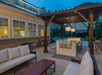 Outdoor patio with wicker furniture, a wooden pergola, a grill, and hanging lights. Building with multiple windows in the background during dusk.