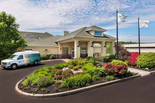 Hotel exterior with a green roof, landscaped gardens, and a parking lot. A shuttle van is parked nearby. Two flags are flying next to the building under a partly cloudy sky.