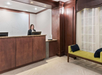 Hotel reception area with a woman standing at the front desk. The room has wood paneling, a patterned chair, and a textured wall.