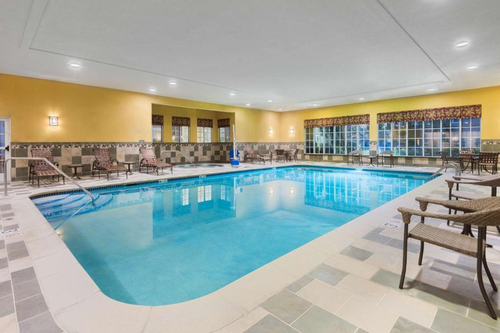 Indoor swimming pool area with clear water, surrounded by lounge chairs and tables. Large windows on two sides allow natural light to enter. Walls are painted yellow with tiled accents.