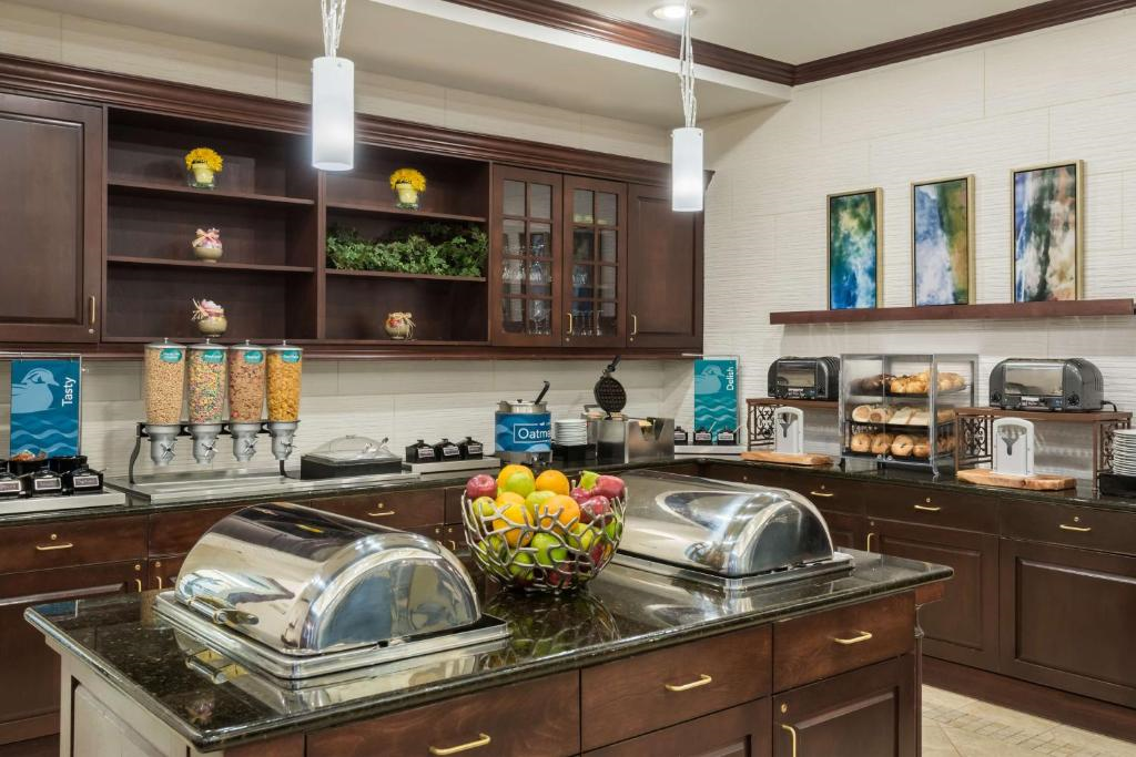Breakfast buffet area with cereal dispensers, bagels, covered trays, a fruit bowl, and toaster. Dark wood cabinetry, framed art on the wall, and pendant lighting overhead.