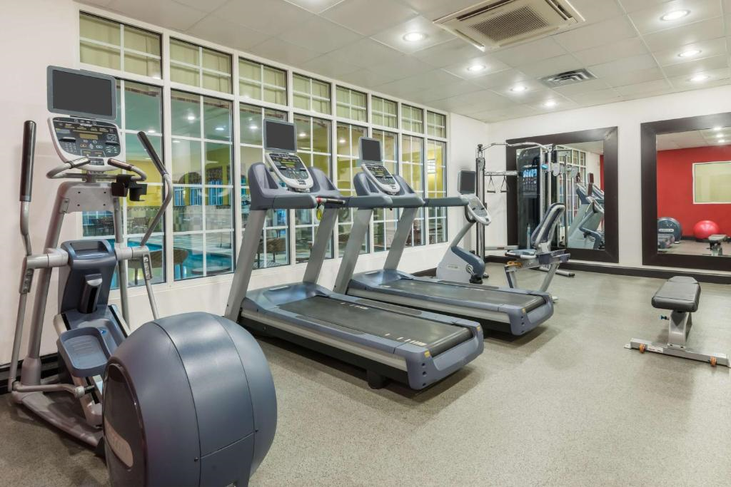 A gym room with three treadmills, exercise bikes, a weight bench, and a large mirror. The space is well-lit with overhead lights and has a windowed wall.