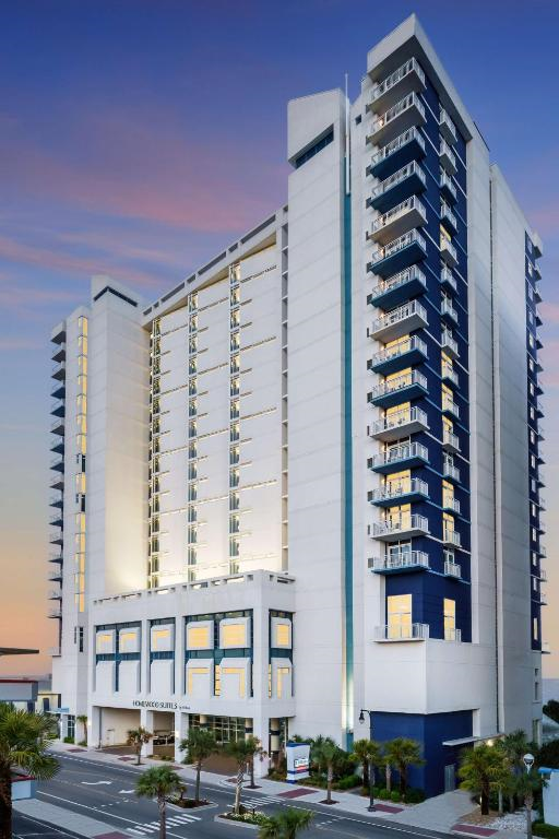 A modern high-rise hotel building with balconies, illuminated at dusk, and palm trees lining the street in front.