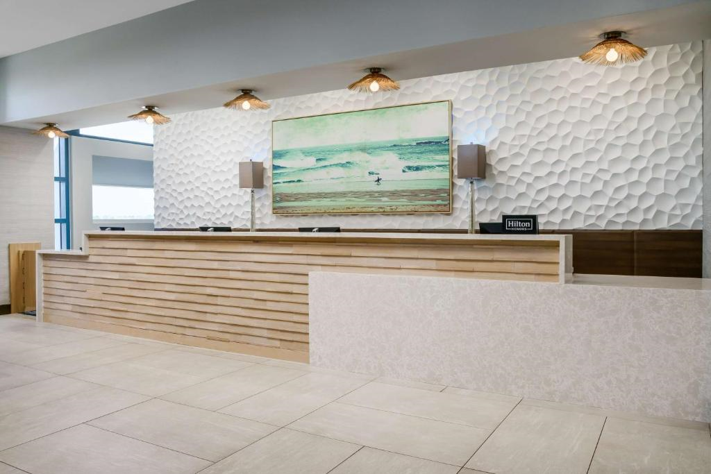 A hotel front desk area with a textured white wall, a wooden counter, overhead lights, and a large painting of ocean waves above the desk.