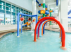 Indoor children's water play area with shallow pool, colorful arches spraying water, water buckets tipping, and a multi-level play structure in the background.