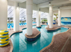 Indoor lazy river pool with large windows, white pillars, stacked yellow float tubes, blue lounge chairs, and a painted mural on the wall.