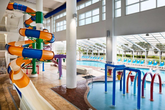 Indoor aquatic center with a twisty water slide, splash play area, and large lap pool, surrounded by lounge chairs and large windows.