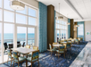 A bright dining area with large floor-to-ceiling windows, blue patterned carpet, and tables and chairs overlooking the ocean.