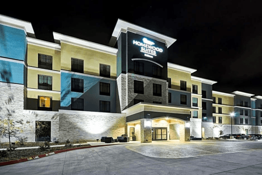 The blue, gray, and yellow front exterior of the Homewood Suites by Hilton New Braunfels with lights casting shadows on it at night.