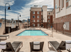 Outdoor pool with lounge chairs and umbrellas, bordered by a metal fence. Residential buildings surround the area. Sky is partly cloudy.