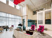 Spacious hotel lobby with high ceilings, modern furniture, pink and purple chairs, a grand piano, and large windows with natural light.