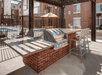 Outdoor patio with a brick grill station, two stainless steel grills, countertop seating with high chairs, and a view of a swimming pool surrounded by residential buildings.