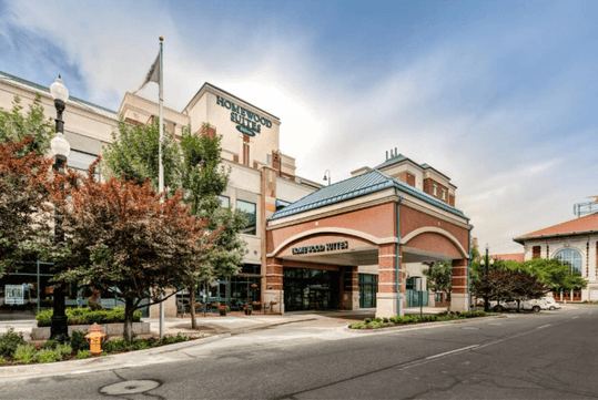 Street view of a hotel with "Homewood Suites" signage, an entrance canopy, surrounding trees, and streetlamps under a partly cloudy sky.