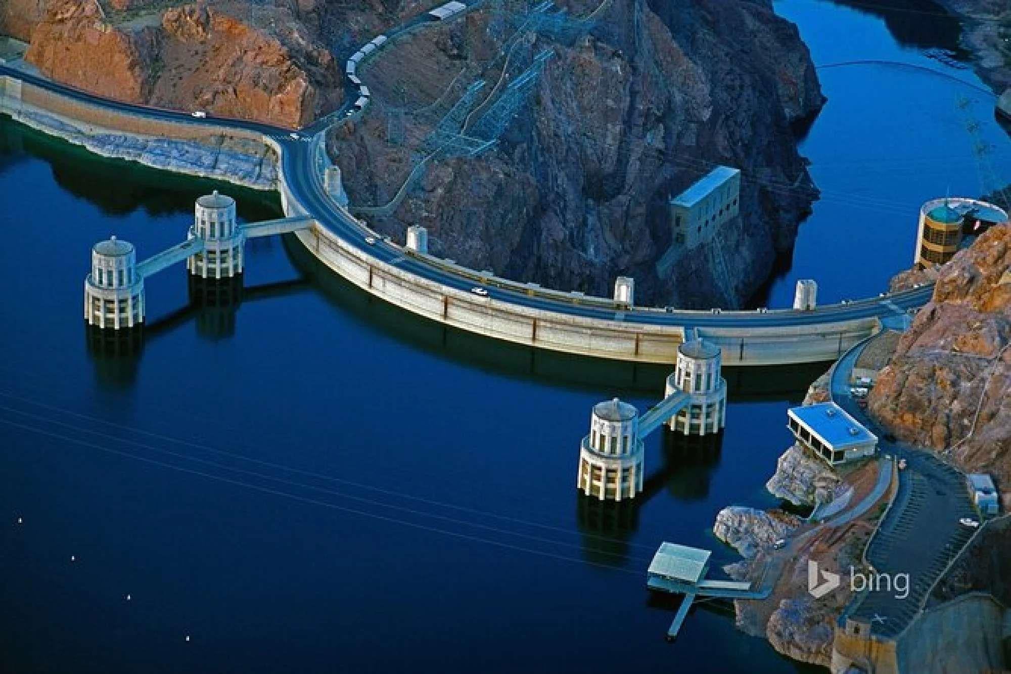 Aerial view of the Hoover Dam spanning the Colorado River, with intake towers, a reservoir on one side, and rugged desert cliffs in the background.