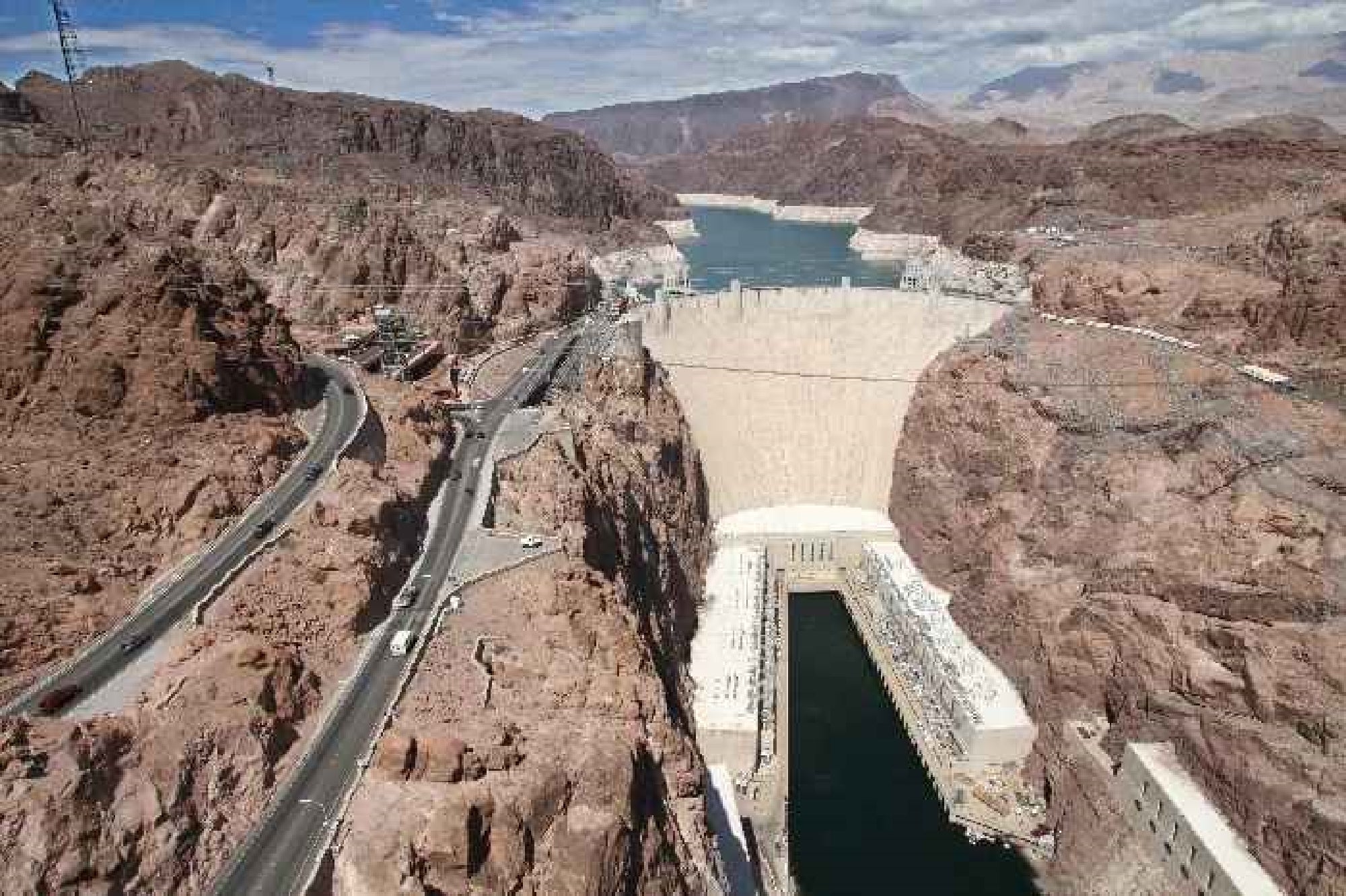 Aerial view of Hoover Dam spanning a river between rocky cliffs, with roads on both sides and mountainous landscape in the background.