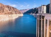 View of a large dam structure in the foreground with a deep blue reservoir surrounded by rocky, arid mountains under a clear blue sky.