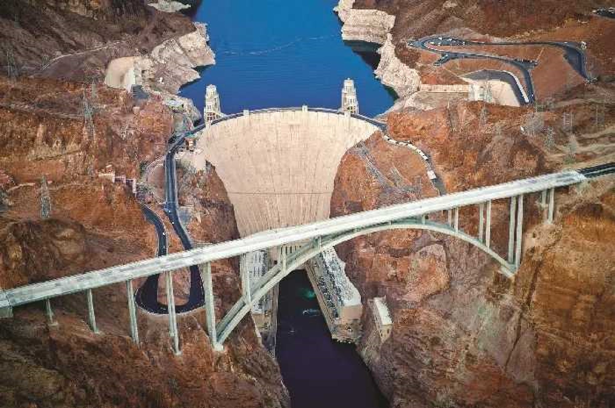 Aerial view of a large concrete dam set between rocky cliffs with a curving bridge spanning the river below.