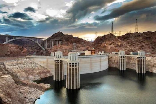 Hoover Dam in a rocky desert landscape, with intake towers rising from the reservoir and a bridge spanning the canyon in the background under a cloudy sky.