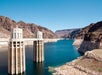 Two intake towers rise from a large reservoir surrounded by rocky, arid cliffs under a clear blue sky.