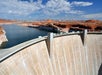 A large concrete dam curves across a river, holding back a reservoir surrounded by red rock formations under a partly cloudy sky.