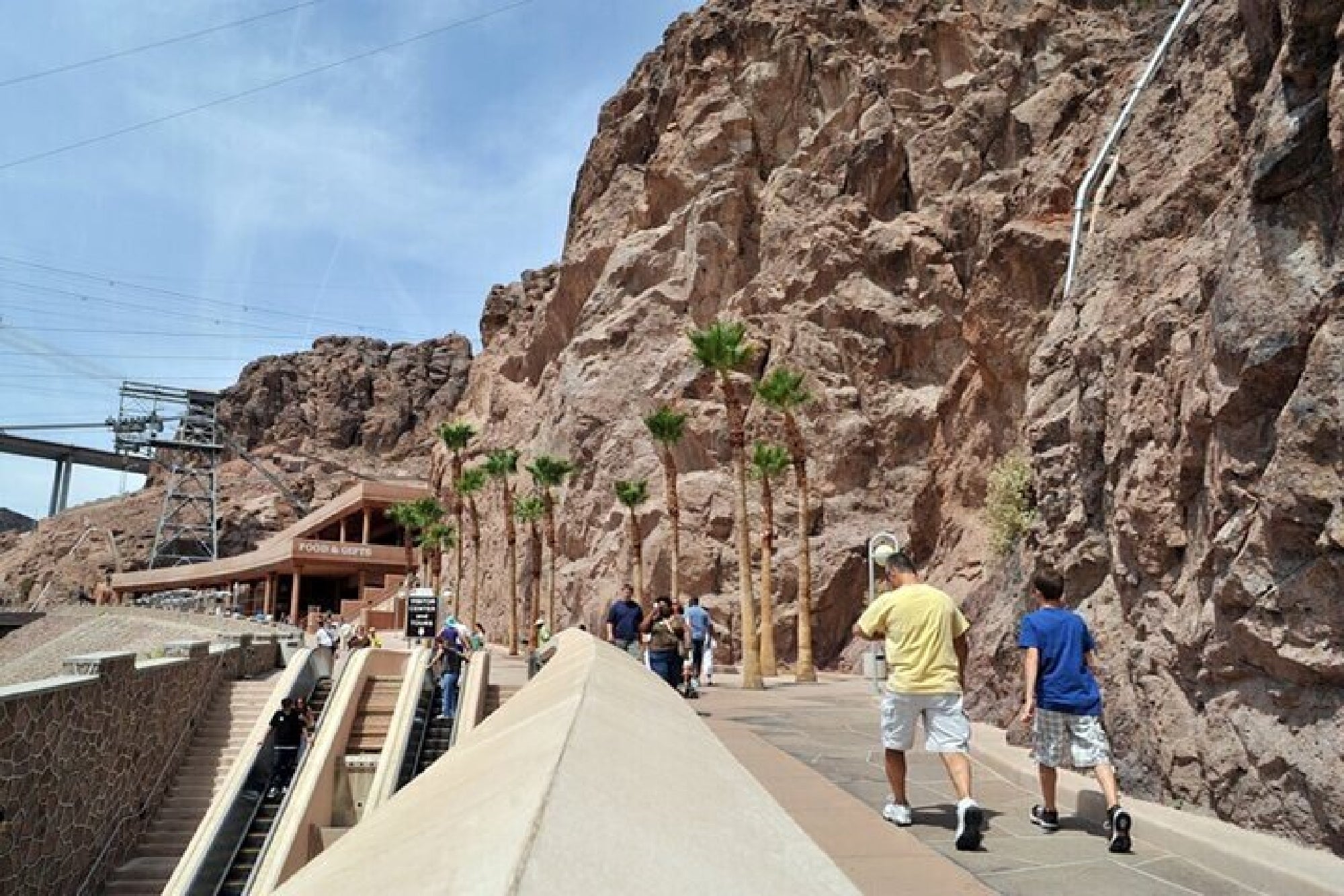 People walk along a paved pathway lined with palm trees next to a rocky cliff near the Hoover Dam Visitor Center under a clear sky.