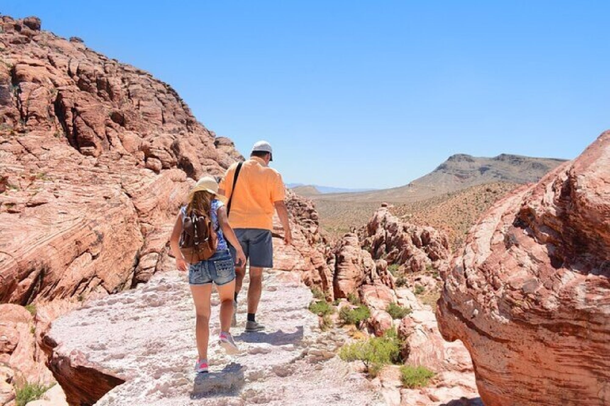 Two people wearing hats and backpacks walk on a rocky trail through a red rock desert landscape under a clear blue sky.