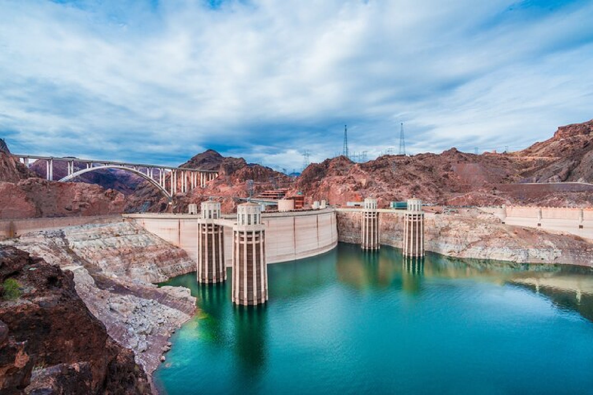 The image shows Hoover Dam spanning a deep canyon with turquoise water below, surrounded by rocky terrain and a bridge in the background under a partly cloudy sky.